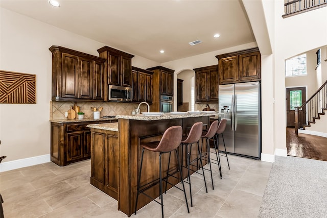 4312 Oxbow Dr, Mckinney, TX 75072 - kitchen featuring appliances with stainless steel finishes, arched walkways, a center island with sink, recessed lighting, and dark brown cabinets