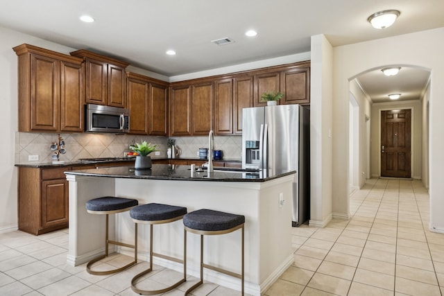 8100 Delaware Dr, Mckinney, TX 75070 - kitchen featuring stainless steel appliances, arched walkways, light tile patterned floors, a kitchen island with sink, and decorative backsplash