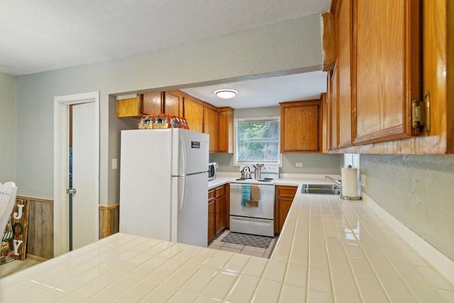 813 Pine St, Teague, TX 75860 - kitchen with white appliances, tile counters, brown cabinetry, wainscoting, and light tile patterned floors