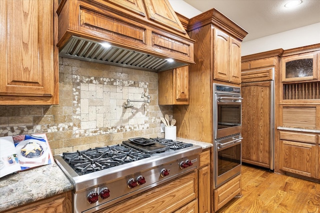 2702 Hidden Trails Rd, Royse City, TX 75189 - kitchen featuring custom exhaust hood, stainless steel appliances, tasteful backsplash, light stone countertops, and brown cabinets