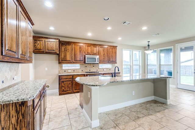 302 Shire Ln, Celina, TX 75009 - kitchen with stainless steel microwave, tasteful backsplash, light stone counters, light tile patterned flooring, and a kitchen island with sink
