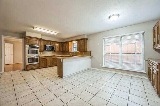 2413 Golden Willow Ln, Richardson, TX 75082 - kitchen featuring appliances with stainless steel finishes, a peninsula, light countertops, brown cabinetry, and light tile patterned floors