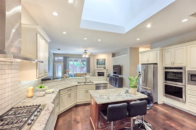 5513 Hill Haven Ct, Frisco, TX 75035 - kitchen with stainless steel appliances, wall chimney exhaust hood, a fireplace, light stone countertops, and a peninsula