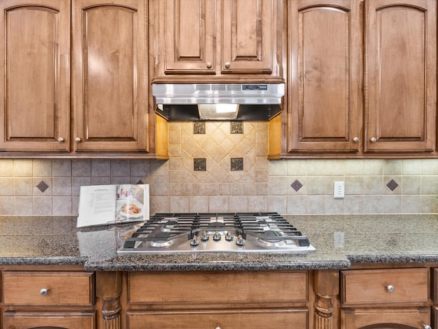 3241 Mindy Ln, Midlothian, TX 76065 - kitchen with under cabinet range hood, stainless steel gas stovetop, tasteful backsplash, brown cabinetry, and dark stone countertops