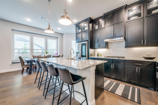 3418 River Trl, Frisco, TX 75034 - kitchen featuring appliances with stainless steel finishes, under cabinet range hood, decorative backsplash, light stone counters, and light wood-type flooring