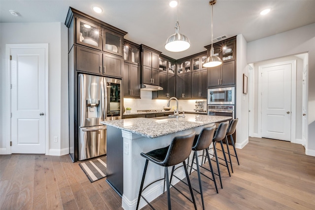 3418 River Trl, Frisco, TX 75034 - kitchen with stainless steel appliances, dark brown cabinets, light wood finished floors, light stone counters, and under cabinet range hood