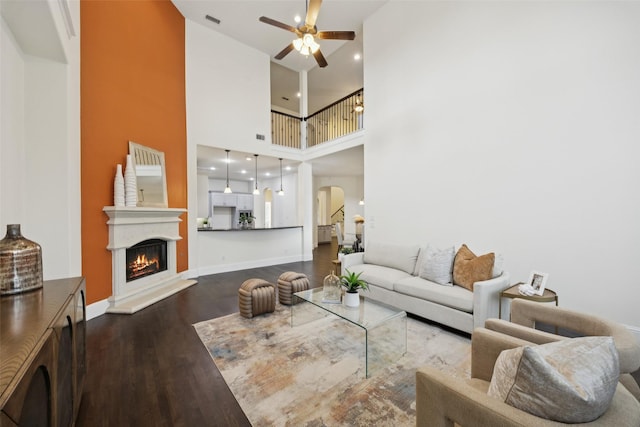 6873 Martel Pl, Frisco, TX 75035 - living area with arched walkways, a ceiling fan, dark wood-style flooring, a warm lit fireplace, and a towering ceiling