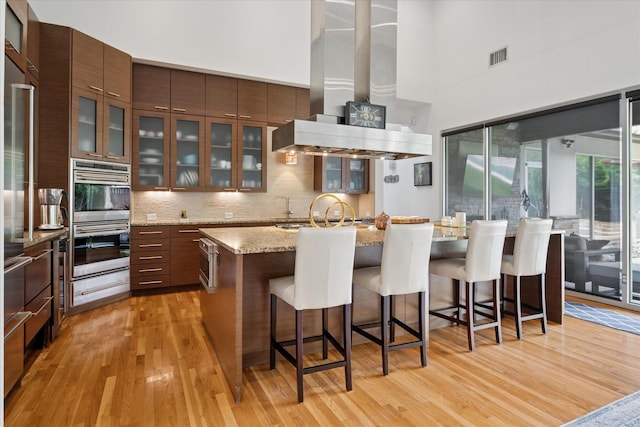 613 N Bailey Ave, Fort Worth, TX 76107 - kitchen featuring light wood-style floors, stainless steel appliances, a towering ceiling, decorative backsplash, and a kitchen bar