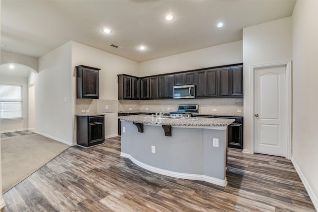 1304 Meridian Dr, Forney, TX 75126 - kitchen featuring appliances with stainless steel finishes, a kitchen island with sink, dark wood-style floors, light stone countertops, and decorative backsplash