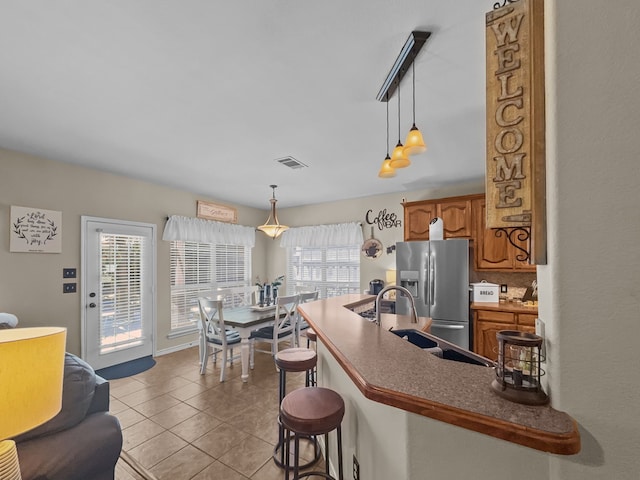 8600 La Quinta Ln, Mckinney, TX 75070 - kitchen featuring stainless steel refrigerator with ice dispenser, brown cabinetry, a kitchen breakfast bar, light tile patterned flooring, and a peninsula