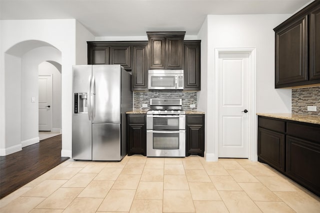 1020 Perennial Ln, Little Elm, TX 75068 - kitchen featuring stainless steel appliances, tasteful backsplash, dark brown cabinets, light stone counters, and arched walkways