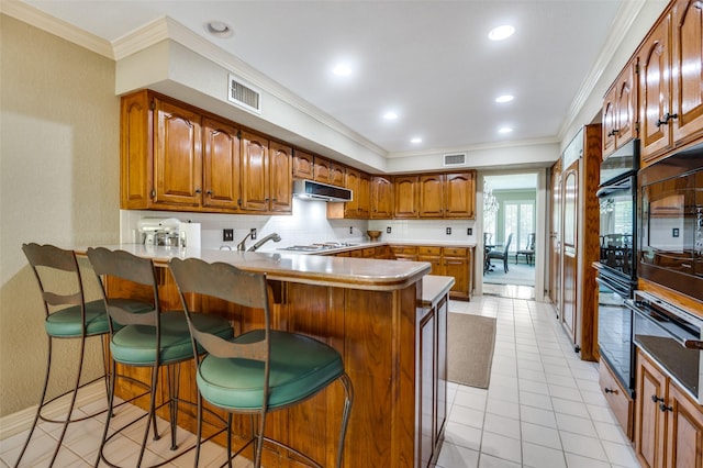 100 La Paz Ct, Irving, TX 75062 - kitchen with under cabinet range hood, ornamental molding, a peninsula, brown cabinets, and tasteful backsplash