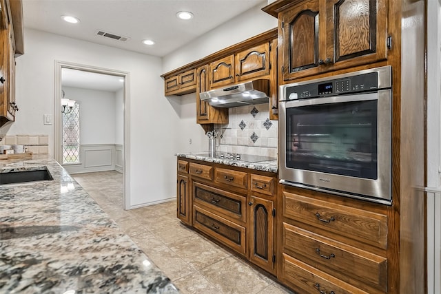 2526 Axminster Dr, Grand Prairie, TX 75050 - kitchen with stainless steel oven, under cabinet range hood, light stone counters, a wainscoted wall, and tasteful backsplash