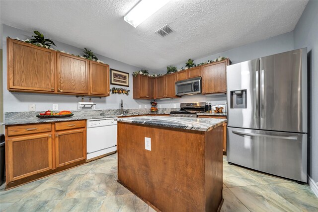 1032 Hampton Dr, Forney, TX 75126 - kitchen featuring appliances with stainless steel finishes, a textured ceiling, a kitchen island, brown cabinetry, and light stone counters