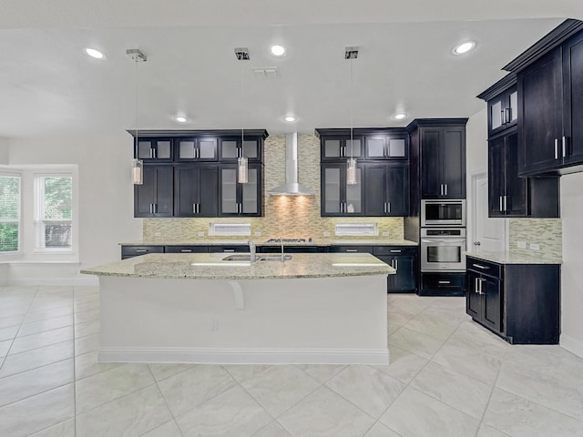 12815 Lourdes Ln, Frisco, TX 75035 - kitchen with wall chimney range hood, stainless steel appliances, a kitchen island with sink, tasteful backsplash, and dark cabinetry