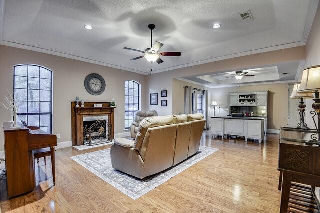 3008 Stone Bluff Ct, Arlington, TX 76017 - living room featuring a ceiling fan, a raised ceiling, crown molding, light wood-style flooring, and a fireplace with flush hearth