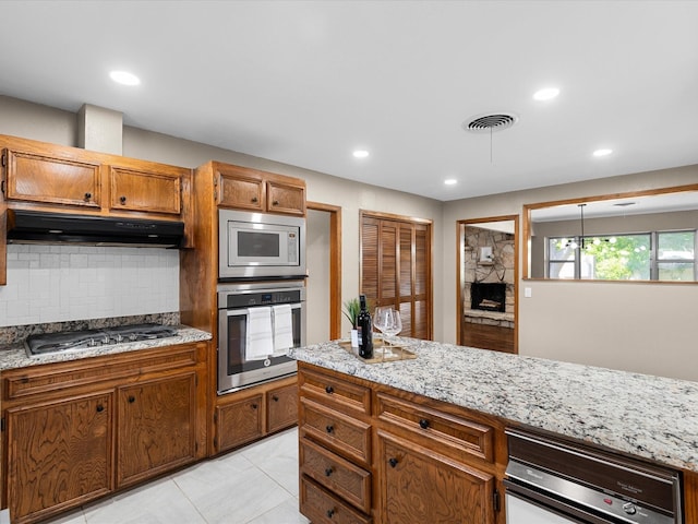 4108 Grand Lake Dr, Lake Worth, TX 76135 - kitchen featuring appliances with stainless steel finishes, under cabinet range hood, tasteful backsplash, brown cabinetry, and light stone countertops