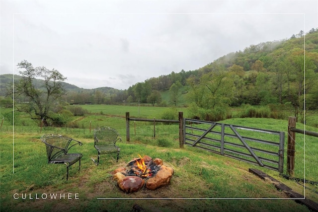view of gate featuring fence, a rural view, a lawn, a view of trees, and an outdoor fire pit