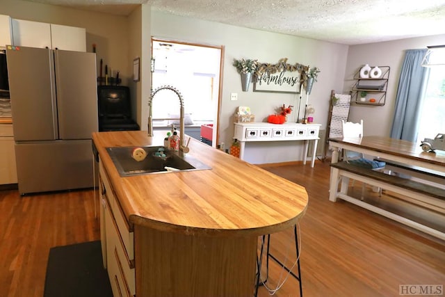 kitchen featuring hardwood / wood-style floors, a textured ceiling, stainless steel fridge, white cabinets, and a kitchen island with sink