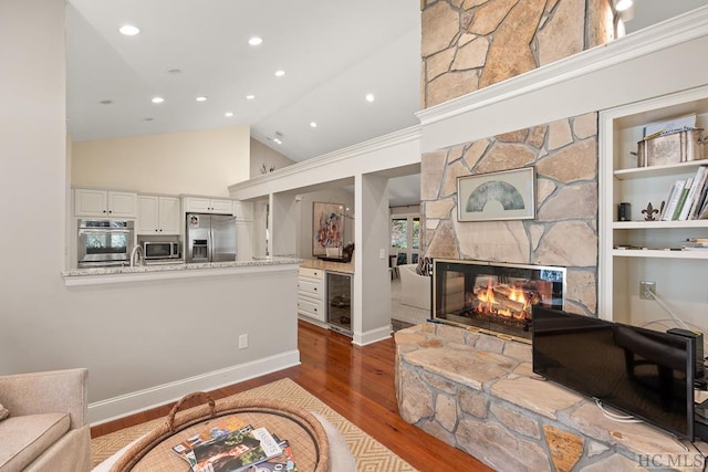 living room featuring a stone fireplace, wood-type flooring, high vaulted ceiling, built in features, and beverage cooler