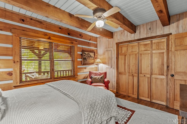 bedroom featuring wood-type flooring, a closet, ceiling fan, wooden walls, and beamed ceiling