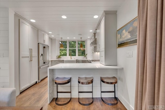 kitchen with high end refrigerator, light wood-type flooring, a kitchen breakfast bar, kitchen peninsula, and wall chimney range hood