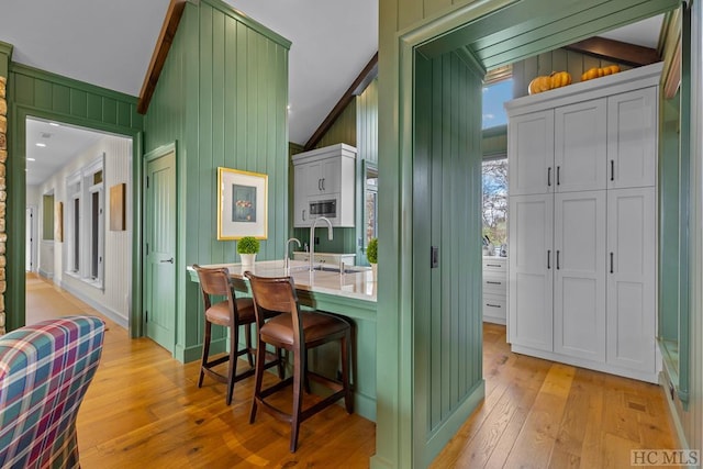 bar with green cabinets, wooden walls, stainless steel microwave, lofted ceiling with beams, and light hardwood / wood-style floors