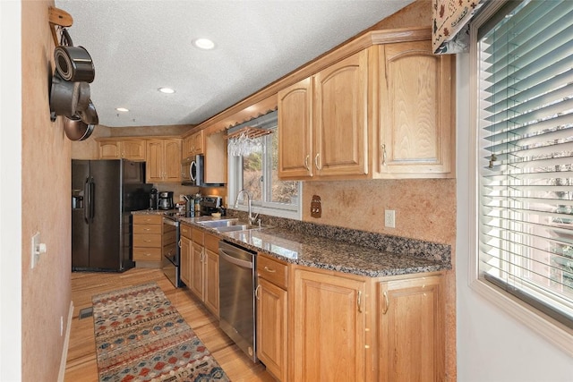 kitchen with appliances with stainless steel finishes, sink, dark stone counters, light brown cabinets, and light wood-type flooring