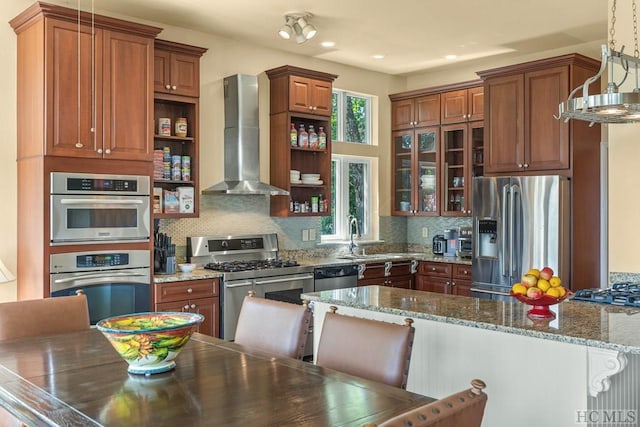 kitchen with hanging light fixtures, wall chimney exhaust hood, stainless steel appliances, dark stone counters, and a kitchen bar