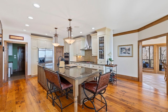 kitchen with pendant lighting, a breakfast bar area, stainless steel appliances, glass insert cabinets, and wall chimney range hood