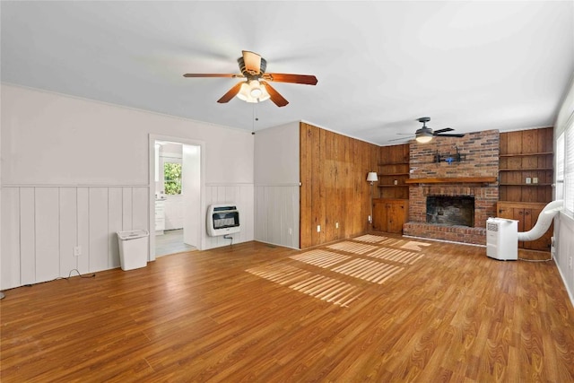 unfurnished living room featuring a fireplace, heating unit, ceiling fan, wood finished floors, and wainscoting