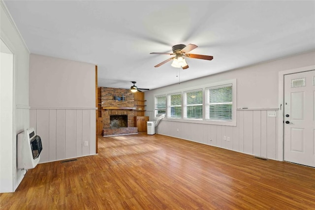 unfurnished living room featuring heating unit, ceiling fan, a brick fireplace, wood finished floors, and a wainscoted wall