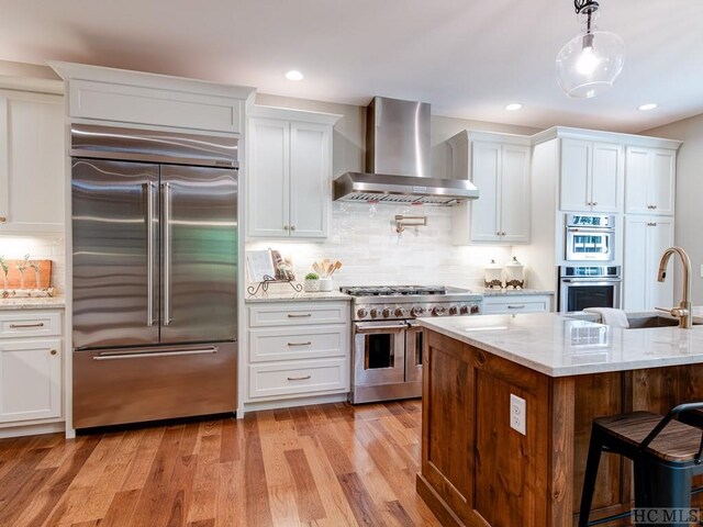 kitchen with high quality appliances, white cabinetry, wall chimney range hood, and decorative light fixtures