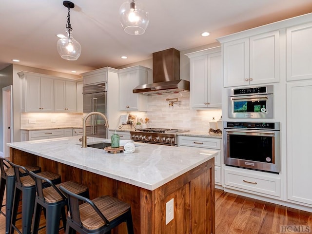 kitchen featuring white cabinetry, a center island with sink, light stone countertops, wall chimney range hood, and high quality appliances