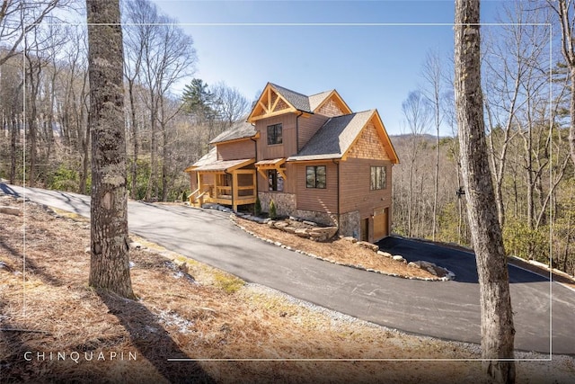 view of front of house with a wooded view, driveway, roof with shingles, an attached garage, and stone siding