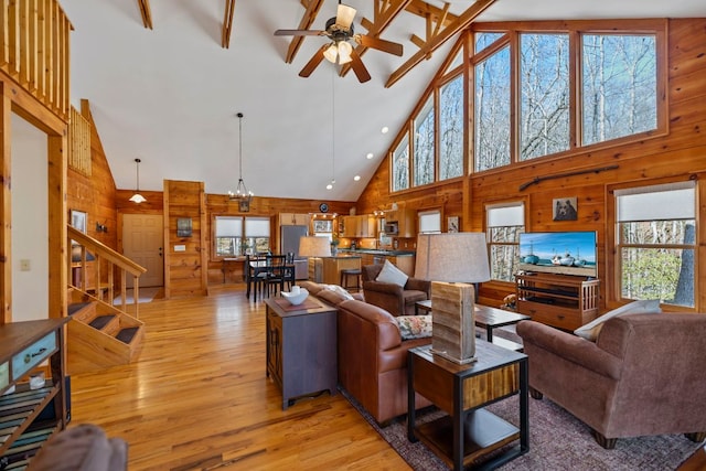 living room with high vaulted ceiling, ceiling fan with notable chandelier, wood walls, light wood-style floors, and stairs