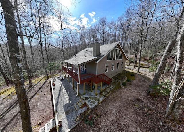 view of side of property with roof with shingles, a wooden deck, a chimney, gravel driveway, and a view of trees