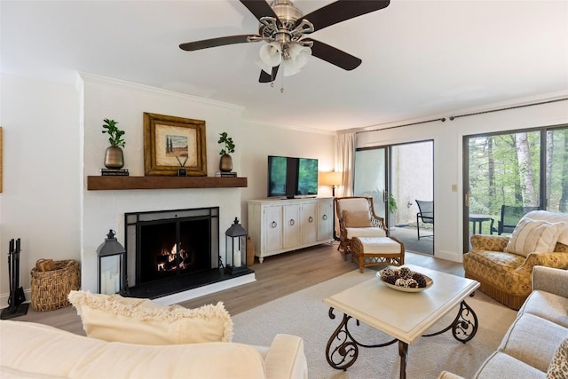 living area with wood finished floors, a ceiling fan, ornamental molding, and a warm lit fireplace