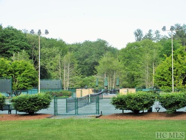 view of tennis court featuring a forest view, a lawn, fence, and a gate