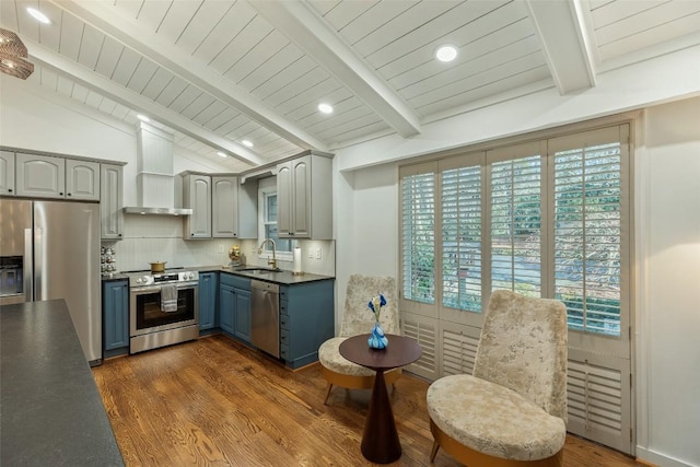 kitchen with blue cabinetry, vaulted ceiling with beams, a sink, appliances with stainless steel finishes, and wall chimney range hood