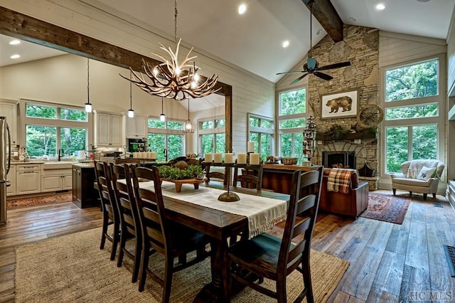 dining room with a stone fireplace, dark hardwood / wood-style floors, ceiling fan with notable chandelier, high vaulted ceiling, and beamed ceiling