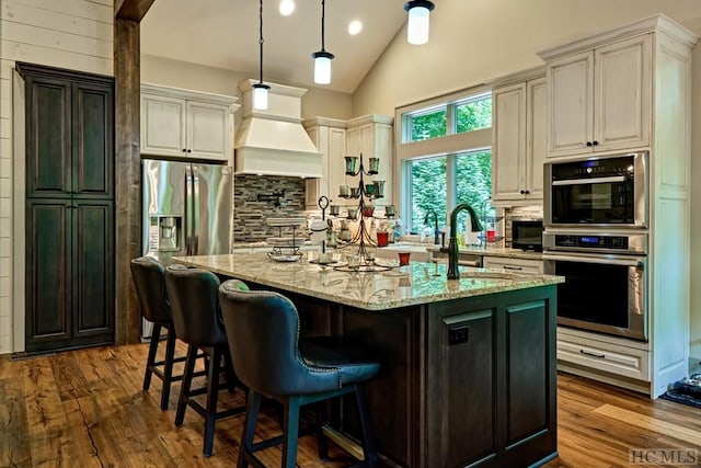 kitchen featuring backsplash, light stone countertops, an island with sink, decorative light fixtures, and custom exhaust hood