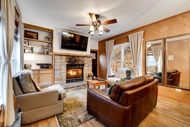 living room featuring built in features, a ceiling fan, light wood-style flooring, a stone fireplace, and wood walls