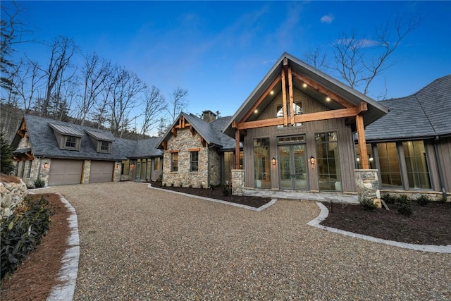 view of front of home featuring an attached garage, stone siding, gravel driveway, and french doors