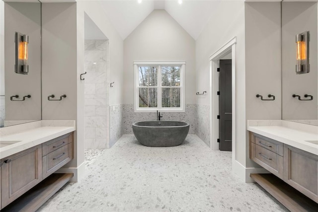 bathroom with a wainscoted wall, tile walls, lofted ceiling, a soaking tub, and two vanities