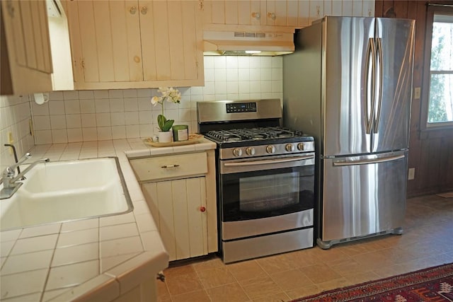 kitchen with under cabinet range hood, a sink, backsplash, appliances with stainless steel finishes, and tile counters