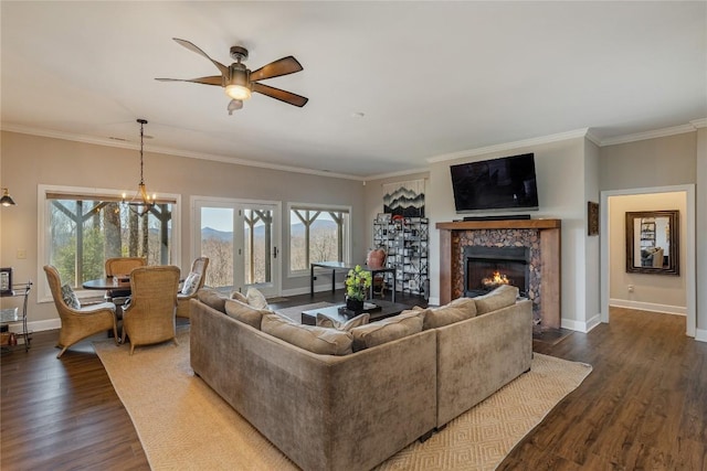 living room featuring dark wood finished floors, a ceiling fan, a chandelier, a stone fireplace, and crown molding