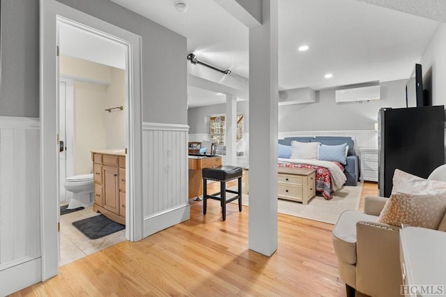 bedroom with light wood-type flooring, stainless steel refrigerator, a wall mounted AC, and ensuite bath