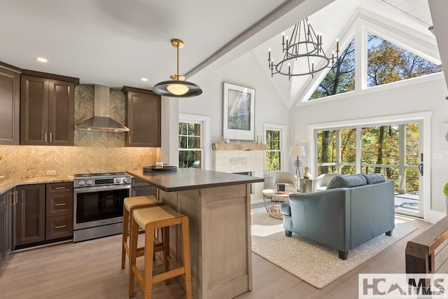 kitchen featuring wall chimney range hood, stainless steel range with electric stovetop, dark brown cabinetry, light wood-style flooring, and high vaulted ceiling