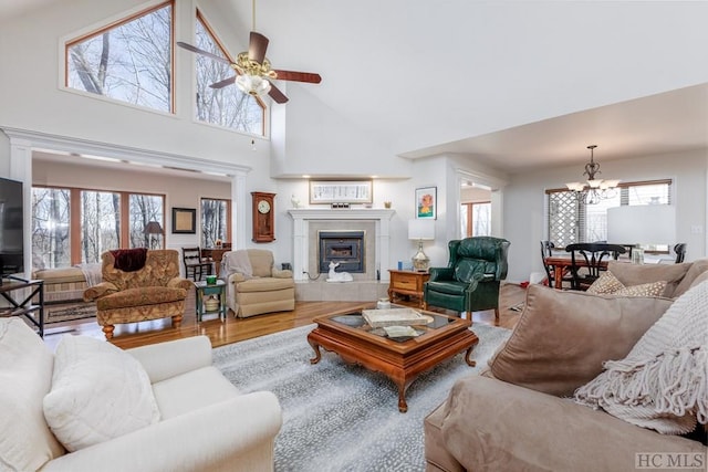 living room with ceiling fan with notable chandelier, wood-type flooring, a tiled fireplace, and high vaulted ceiling